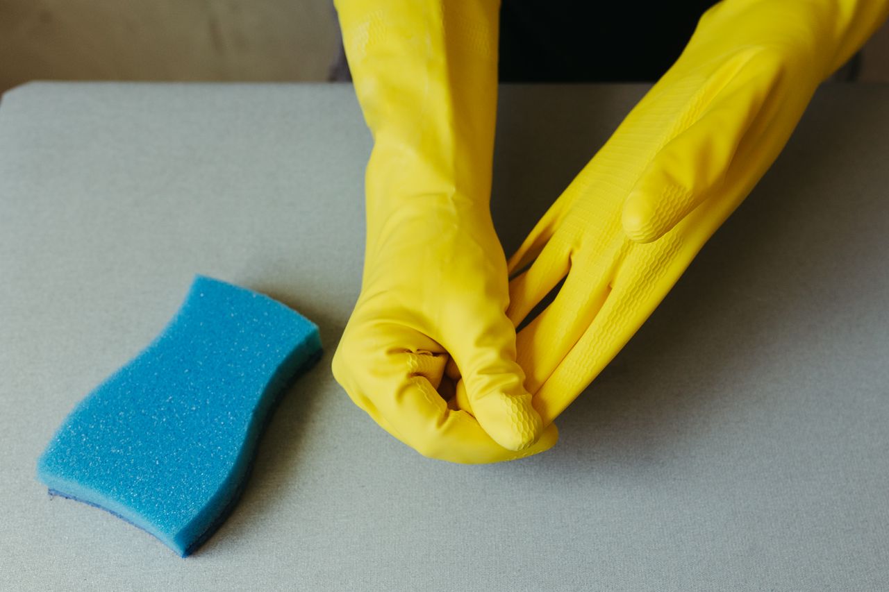 Close-up of hands wearing yellow gloves next to blue sponge on gray surface.