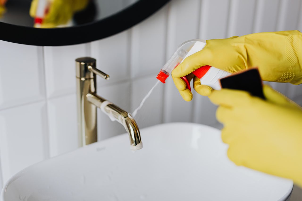 From above an anonymous housekeeper wearing yellow rubber gloves, spraying cleaner onto a chrome bathroom water faucet